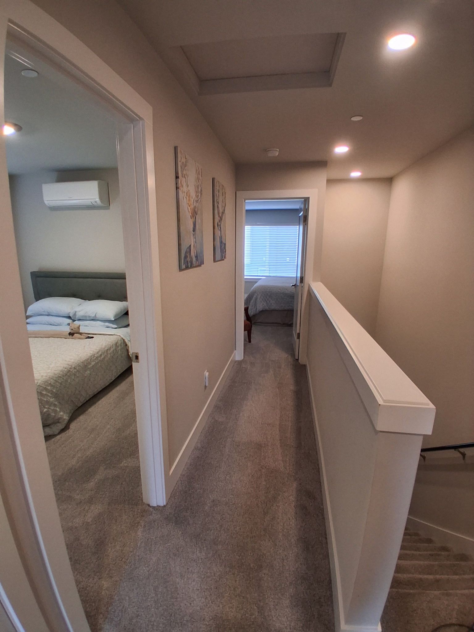 A narrow hallway with carpeted floors, a white railing, and a door leading to a bedroom with a bed and a window.