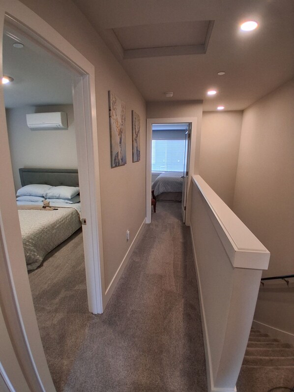 A narrow hallway with carpeted floors, a white railing, and a door leading to a bedroom with a bed and a window.