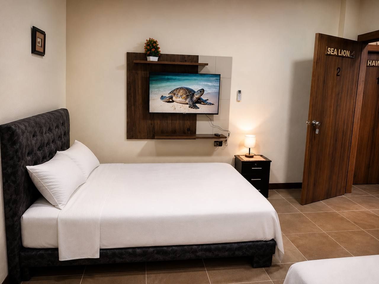 A hotel room with a bed, a television mounted on the wall, a bedside table with a lamp, and a wooden door labeled 'SEA LION'.