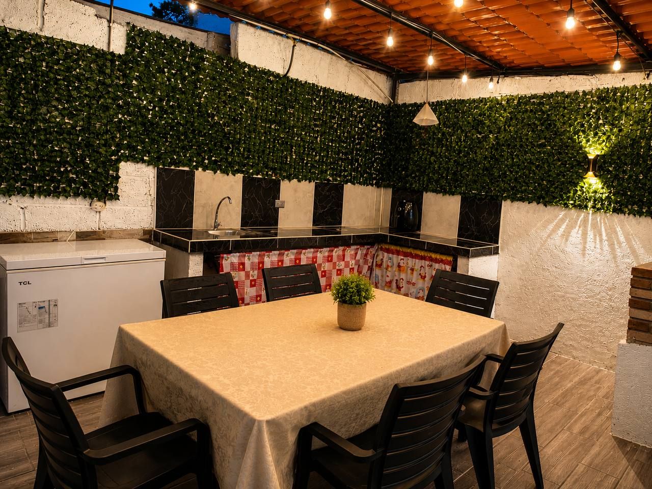 An outdoor dining area with a table and chairs, a refrigerator, and a wall covered with greenery.