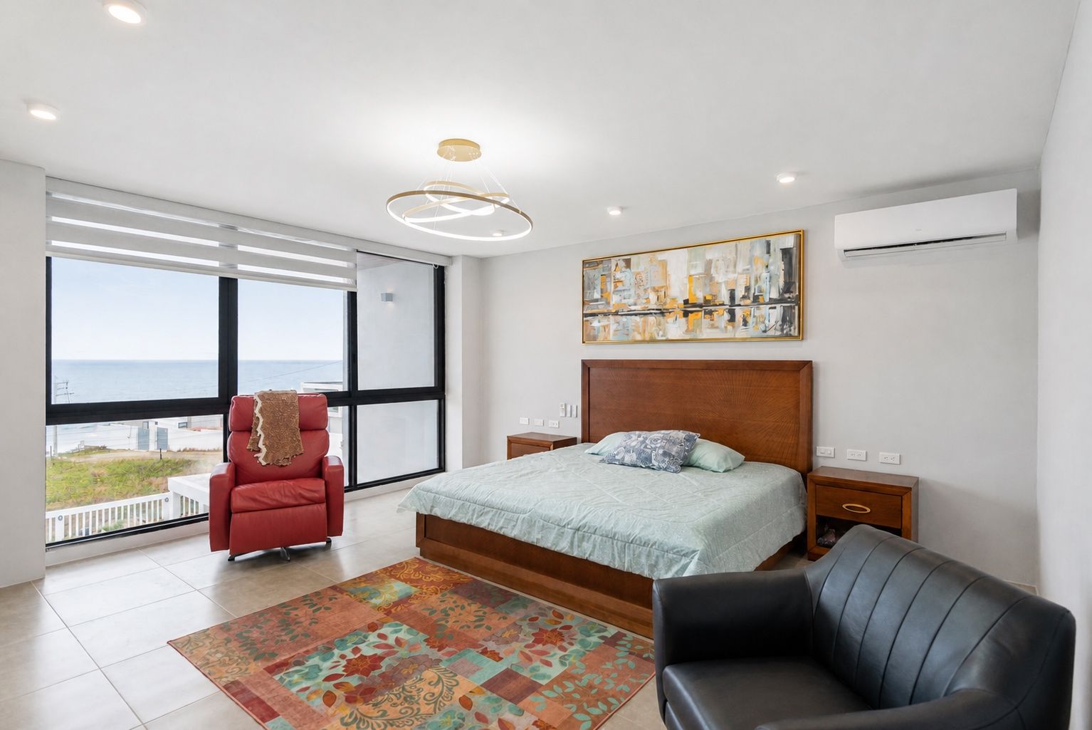 Une chambre moderne avec un grand lit, une chaise rouge, un fauteuil noir et une vue sur l’océan.