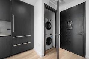 Ein moderner Waschraum mit Waschmaschine und Trockner, einem dunkelholzigen Kleiderschrank und einer Tür mit Notruhinweis.