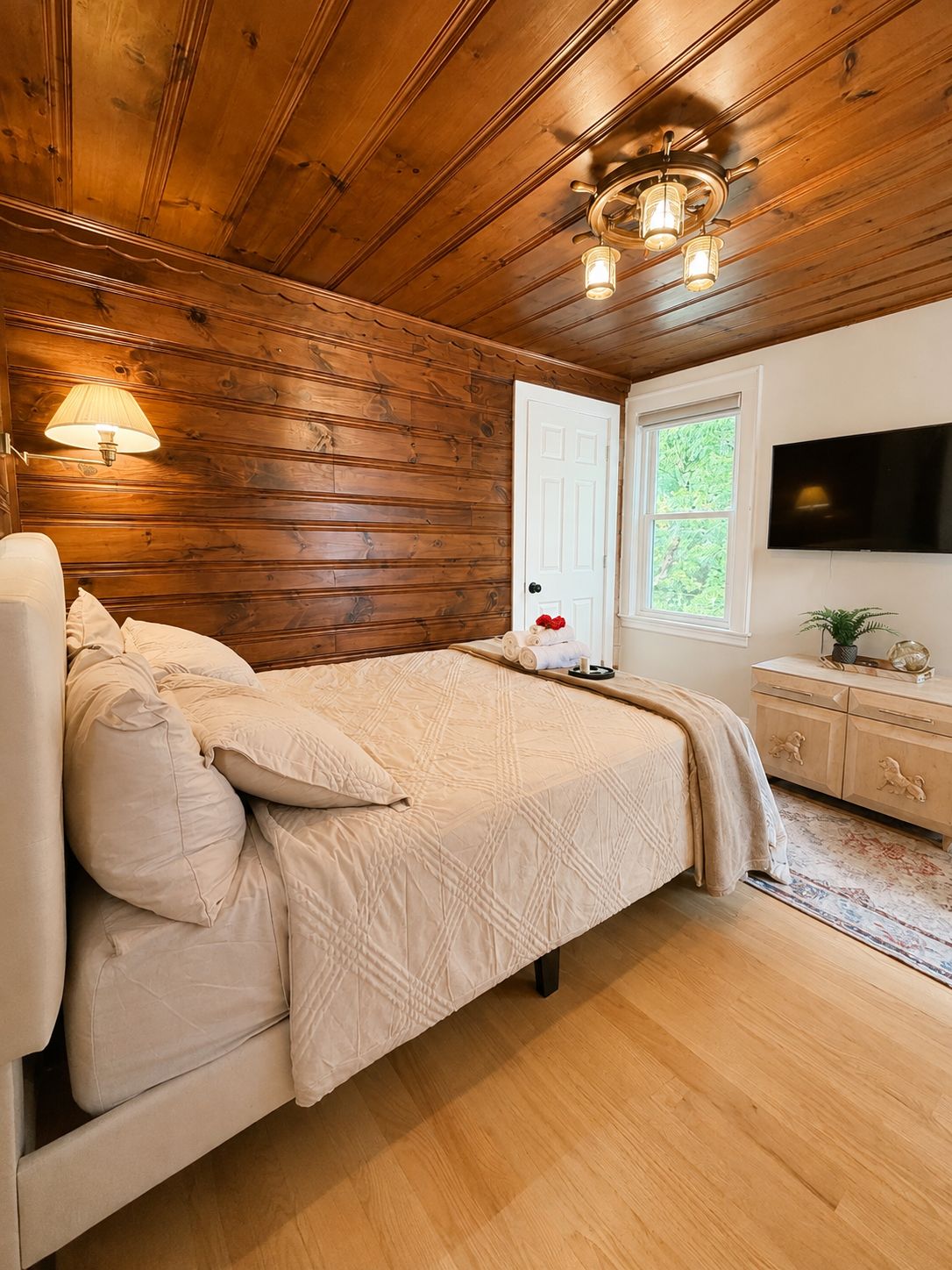 A bedroom with a wooden ceiling, a bed with a beige quilt, a nightstand, a lamp, a TV, and a window with a view of greenery.
