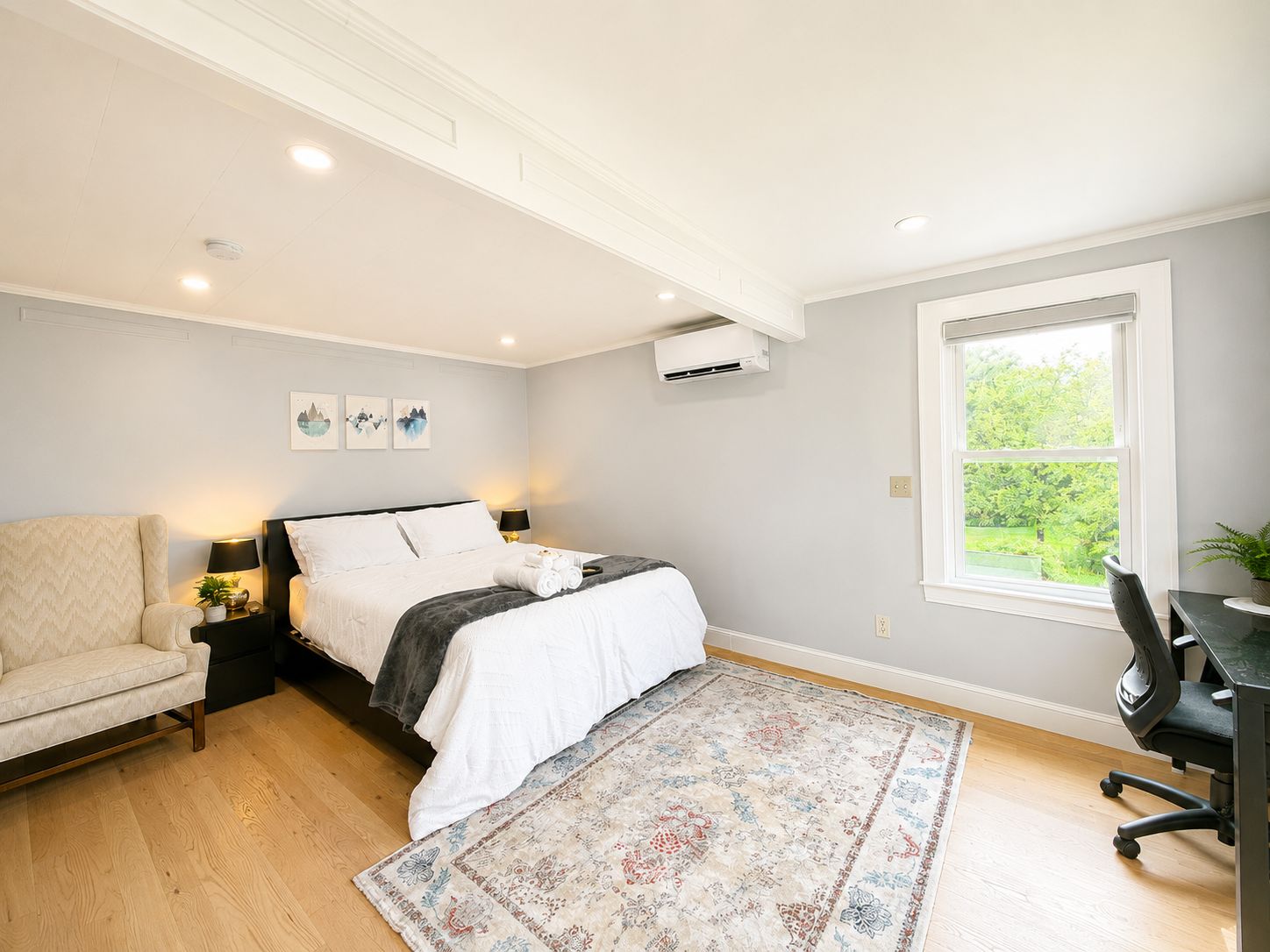 A bedroom with a bed, a chair, a desk, an air conditioning unit, and a window with a view of greenery.