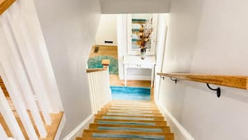 Un escalier avec des marches en bois et une moquette bleue menant à un palier où se trouve une console blanche et un vase de fleurs séchées.