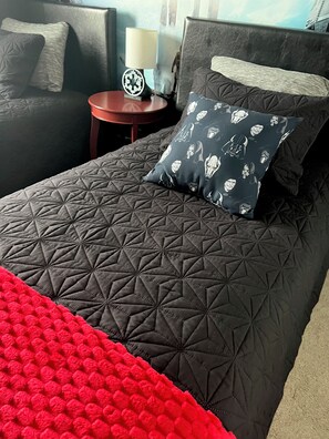 Ein Bett mit einem geometrisch gemusterten Bettbezug, einem Kopfkissen mit Darth-Vader-Motiv und einer roten Decke.