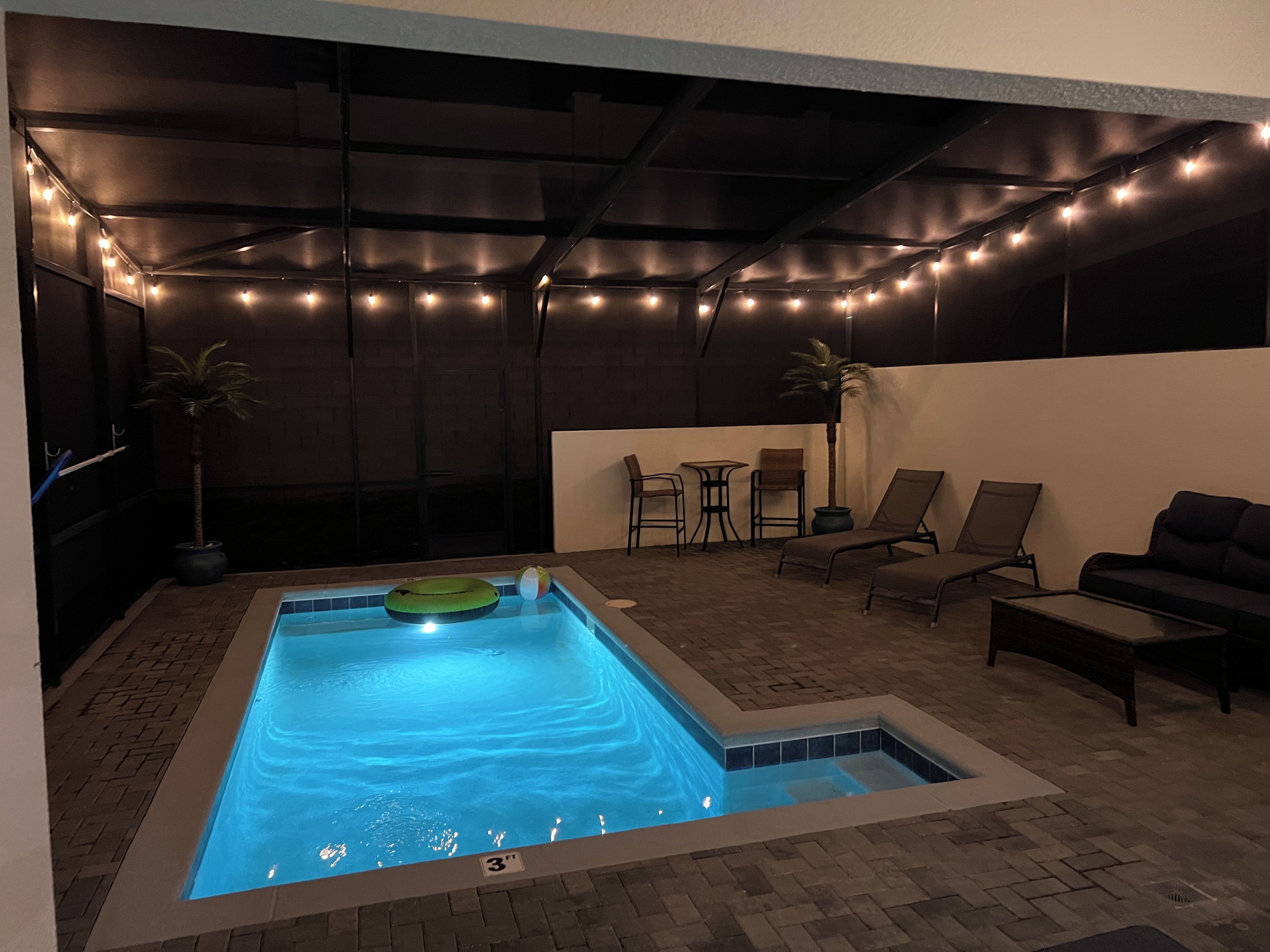 Une piscine rectangulaire dont la profondeur est indiquée par un repère à 3 pieds, entourée de chaises longues et d’une table, le tout illuminé par des guirlandes lumineuses.