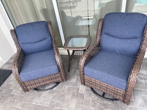 Dos sillones de mimbre con cojines azules y una mesita, ubicados en un patio con baldosas.