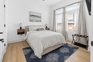 Een slaapkamer met een bed, nachtkastje, wandlamp, raam met gordijnen en uitzicht op het omliggende landschap.
