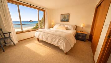 A bedroom with a large window offering a view of the sea, a bed with white bedding, a nightstand with lamps, and a chair by the window.