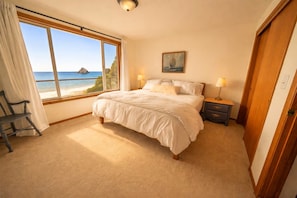 A bedroom with a large window offering a view of the sea, a bed with white bedding, a nightstand with lamps, and a chair by the window.