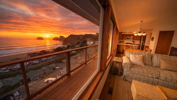 Una habitación con vista al océano al atardecer, una terraza de madera y un rincón de estar acogedor con un sofá y una mesita.