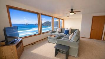 A living room with a large window overlooking the ocean, a television, a sofa, and a ceiling fan.