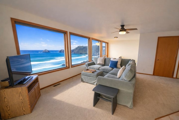 A living room with a large window overlooking the ocean, a television, a sofa, and a ceiling fan.