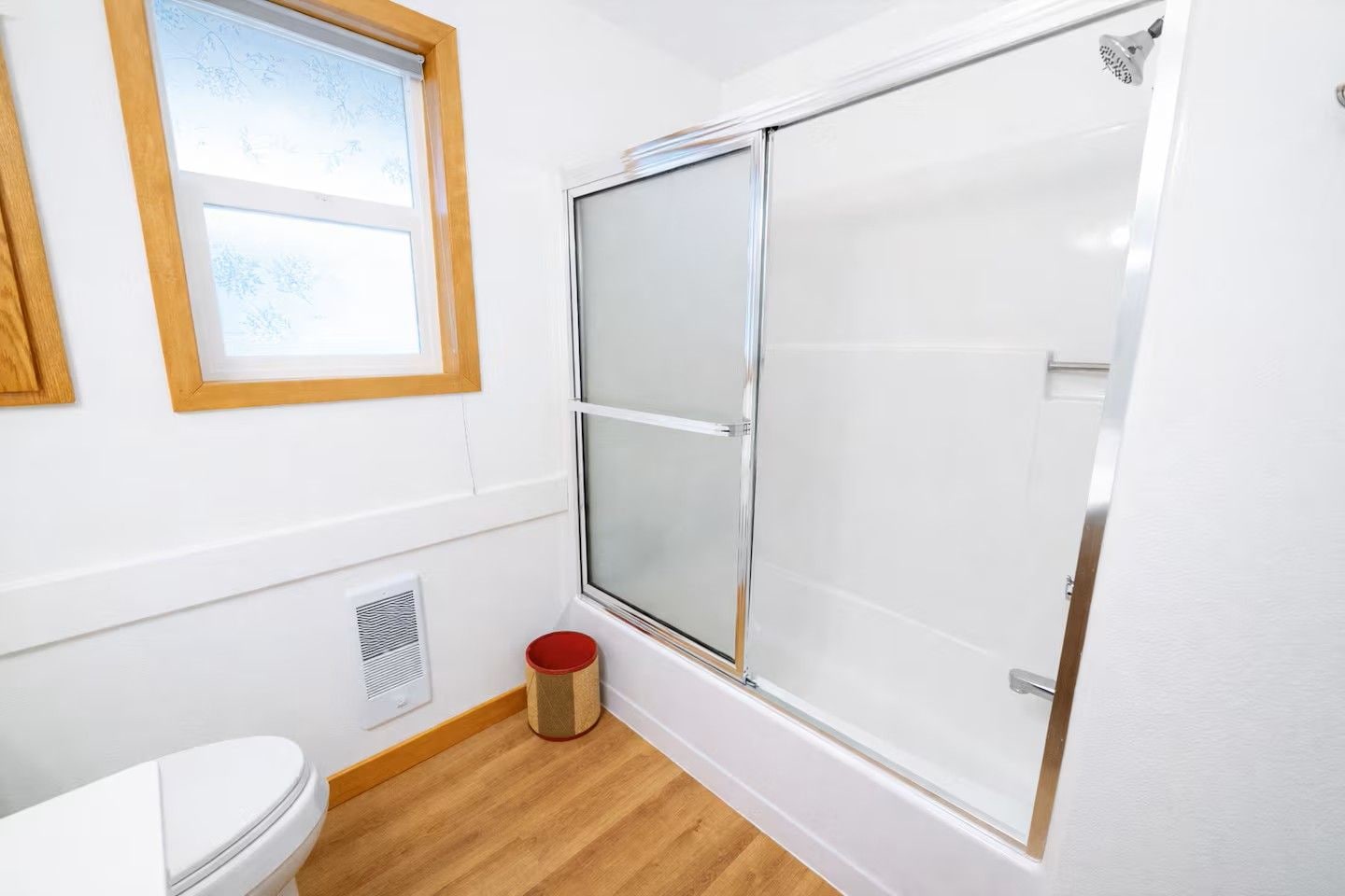 Un baño con inodoro, una ventana, ducha con puerta de cristal y un bote de basura rojo.