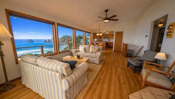 A spacious living room with a large window overlooking a scenic view, a ceiling fan, and a kitchen area visible through an open doorway.