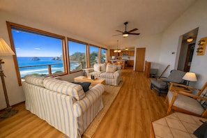 A spacious living room with a large window overlooking a scenic view, a ceiling fan, and a kitchen area visible through an open doorway.