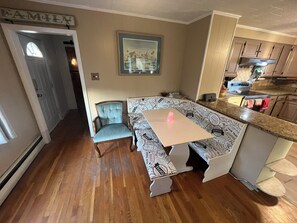 Un comedor con una mesa tipo banca y una silla, una cocina con armarios y electrodomésticos, y un cuadro en la pared.