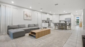 A modern living room with a grey sectional sofa, a wooden coffee table, and a kitchen area with white cabinets and a dining set. - 7BR Villa Solara Resort Pool & Cinema Near Disney (Kissimmee)