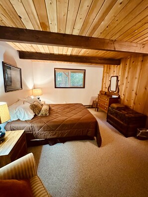 A bedroom with a wooden ceiling, a bed with a brown quilt, a nightstand, a dresser, a mirror, and a window with a view of trees. - The Chalet on Lopez: it's peaceful! (Lopez Island)