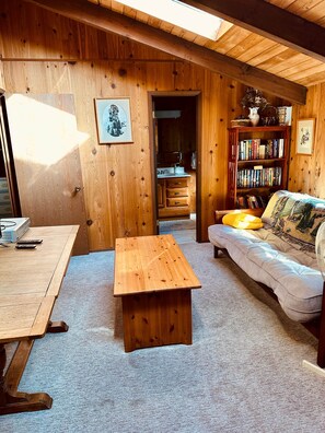 A cozy room with a wooden floor, a sofa, a coffee table, a bookshelf, and a kitchen area visible through an open door. - The Chalet on Lopez: it's peaceful! (Lopez Island)