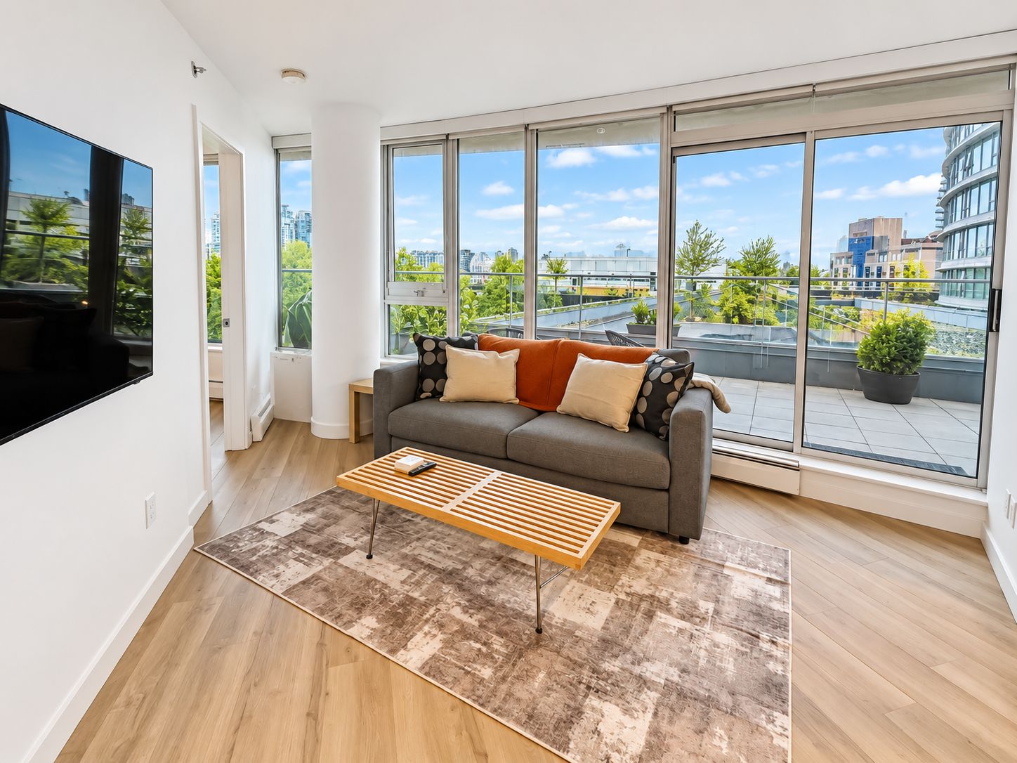 A modern living room with a grey sofa, a wooden coffee table, and large windows offering a city view.