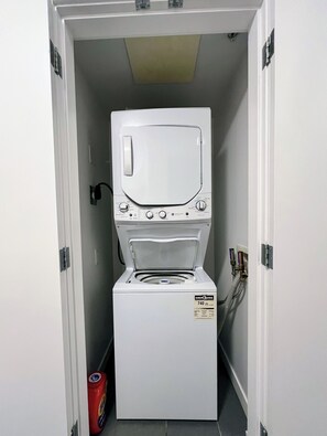 A compact washer and dryer unit in a narrow hallway with a door on one side. - 2BR+Sofabed, 2Bath, Private 500sqft sundeck, EV Parking w/Pool, Sauna, Hot Tub (Vancouver)