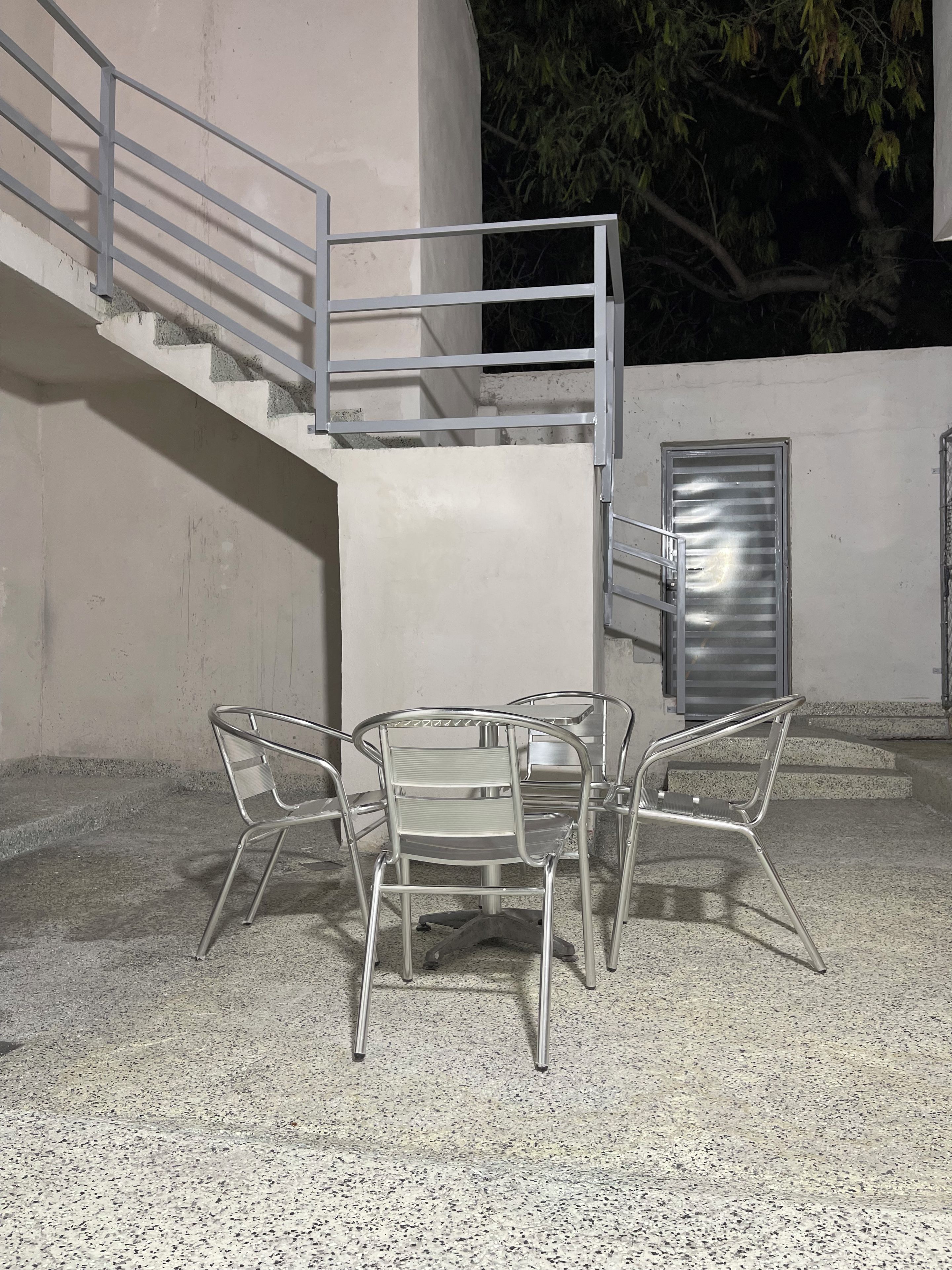 Un conjunto de sillas metálicas y una mesa frente a un edificio con una escalera de concreto y barandilla metálica.