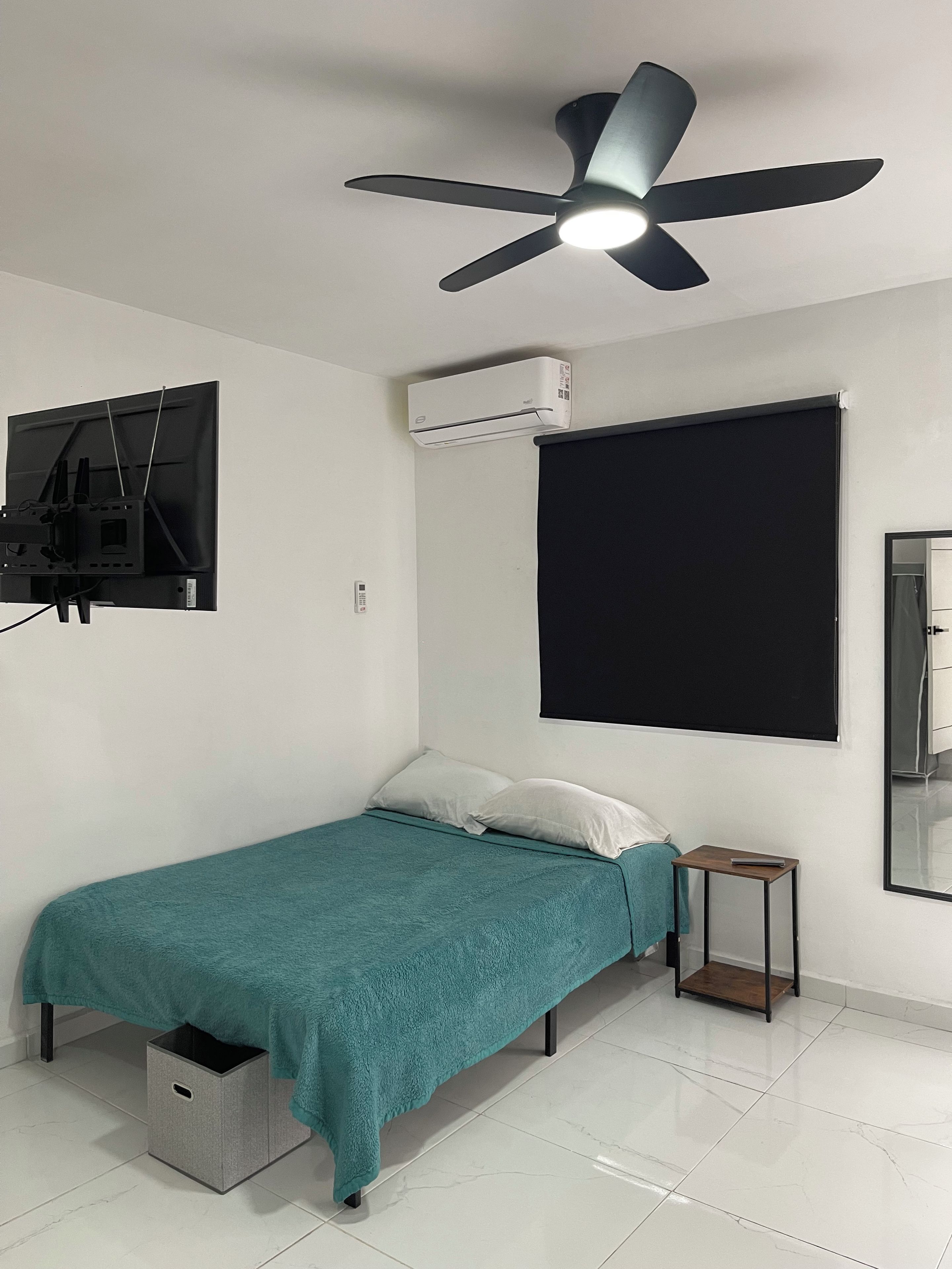 A bedroom with a bed, a ceiling fan, an air conditioning unit, a TV mounted on the wall, and a window with a blind.