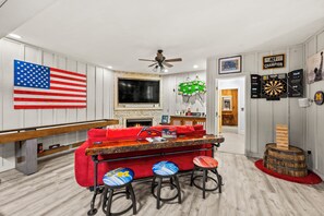 A room with a red couch, two bar stools, a flat-screen TV, a ceiling fan, a dartboard, and a dartboard with a 'Practice Like a Champion' sign. - ** NEW RENTAL** Highlands Heaven | Walk to Town | Game Room | Firepit & Waterfall (Highlands)