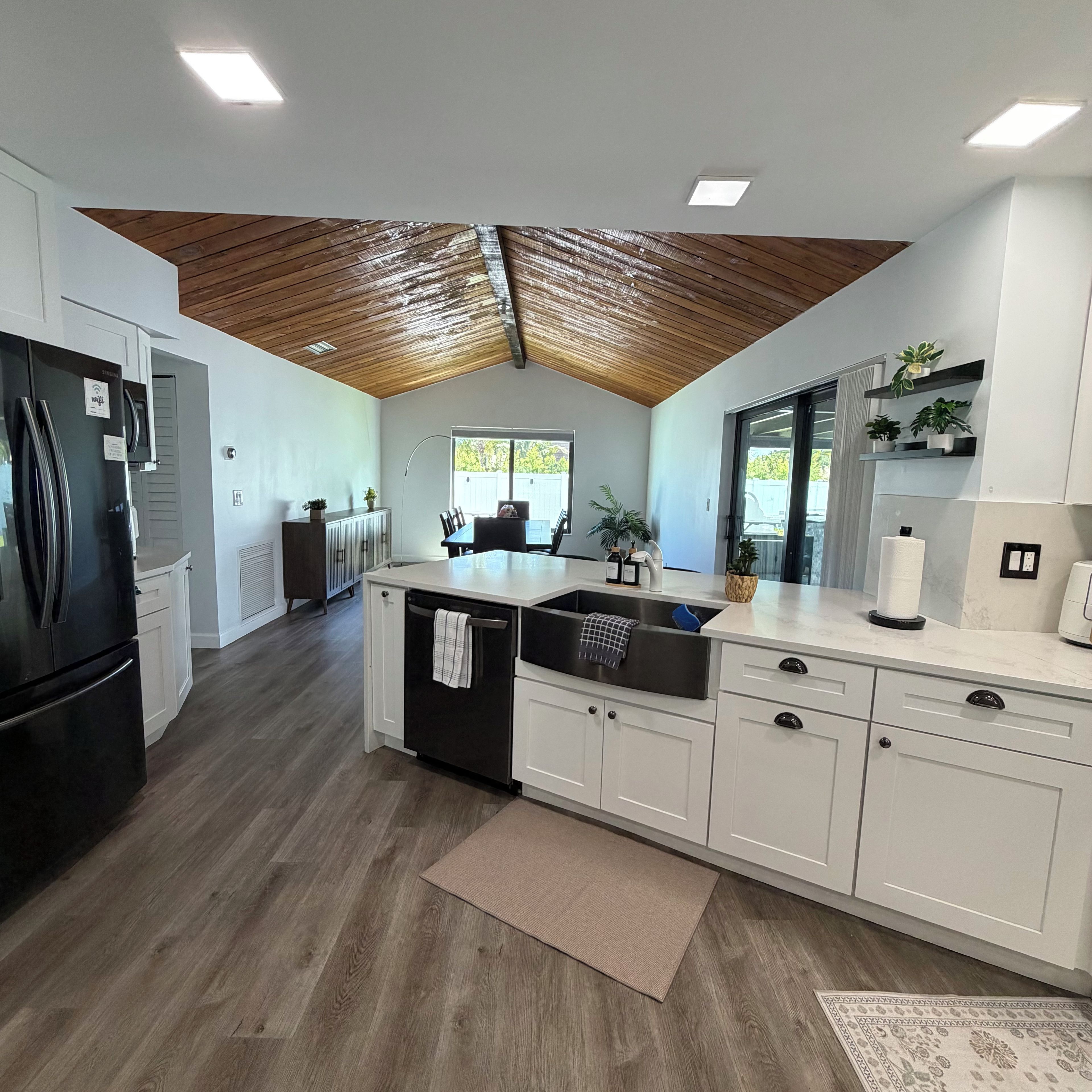 A modern kitchen with a wooden ceiling, white cabinetry, a black refrigerator, and a large window.