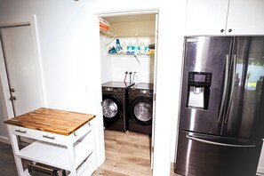 A compact kitchen with a stainless steel refrigerator, a washing machine, and a wooden table with drawers. - Casa Sol: Jacuzzi, Outdoor Kitchen & BBQ in Deerfield Beach (Deerfield Beach)