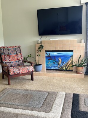 A living room with a flat-screen TV mounted on the wall, a patterned armchair, a fireplace with a painting, and two potted plants.