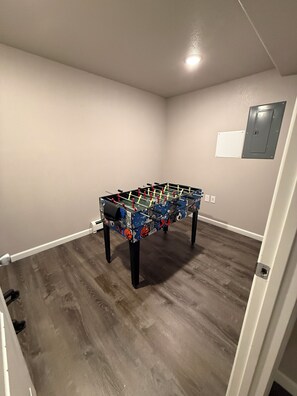 Una habitació amb taula de futbolí, terres de fusta, una porta blanca i un quadre elèctric fixat a la paret.