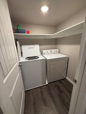 A laundry room with a washer and dryer, wooden flooring, and a shelf above the dryer. - Perfect 2-bedroom apartment in welcoming Loveland for your stay (Loveland)
