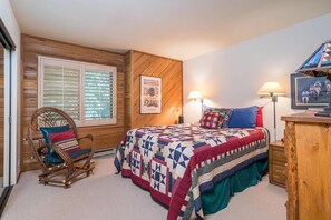 Un dormitorio con piso de madera, una cama con colcha, una silla, una mesita de noche y una lámpara de pared.