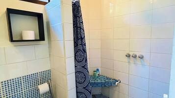 A small bathroom with a shower, toilet, and sink. There is a window above the toilet, a towel rack, and a shelf with toiletries.