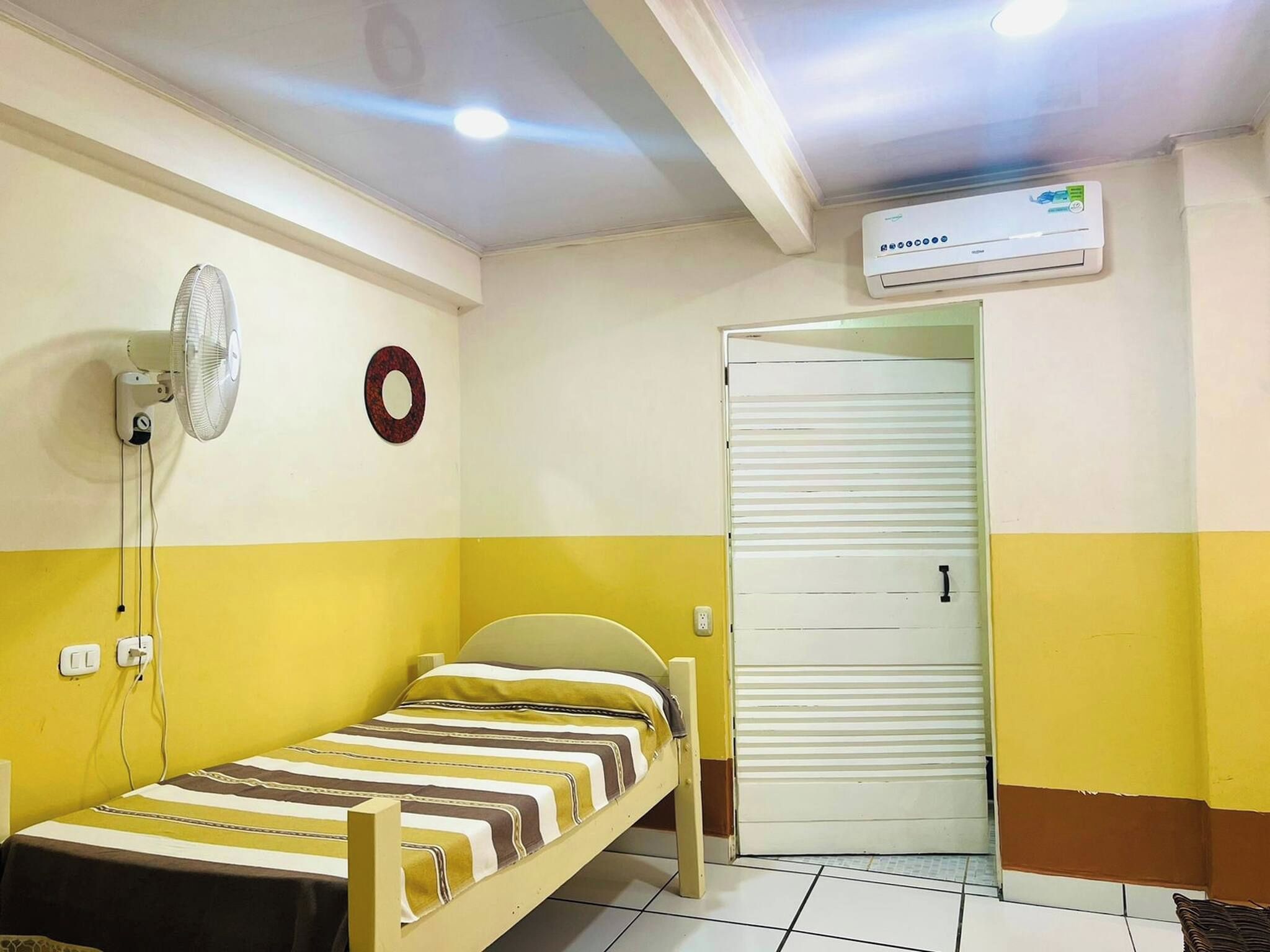 A small room with a bed, a wall-mounted fan, a circular decoration, and an air conditioning unit.