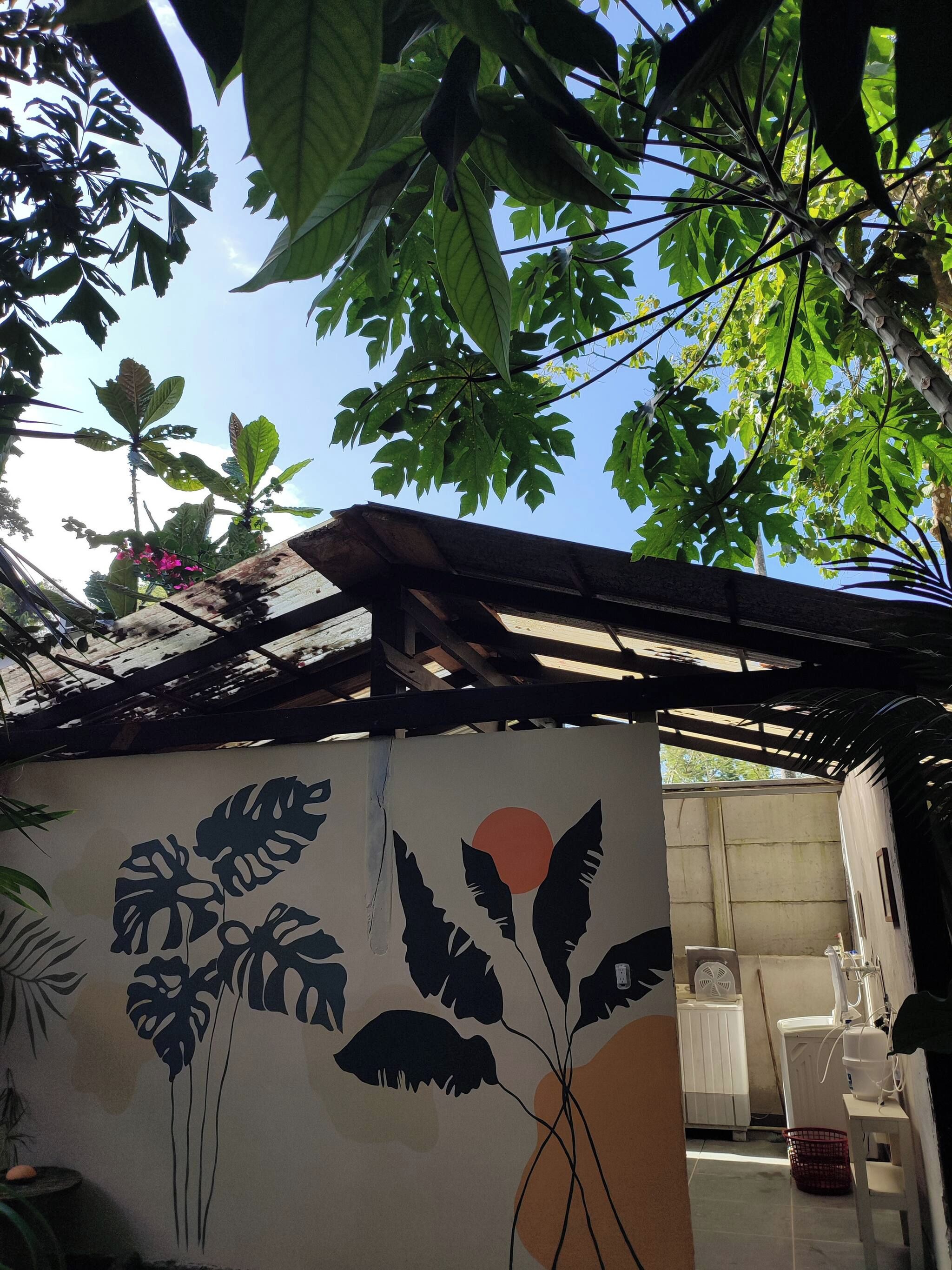 Una habitación con un mural de hojas y un sol, un ventilador y una lavadora.