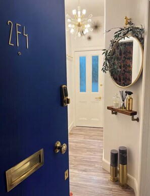 A blue door with a number plate, a hallway leading to a white door with blue accents, a round mirror with a decorative wreath, and two black and gold candle holders. - Luxury Leith Apartment | Stylish Edinburgh Stay (Edinburgh)