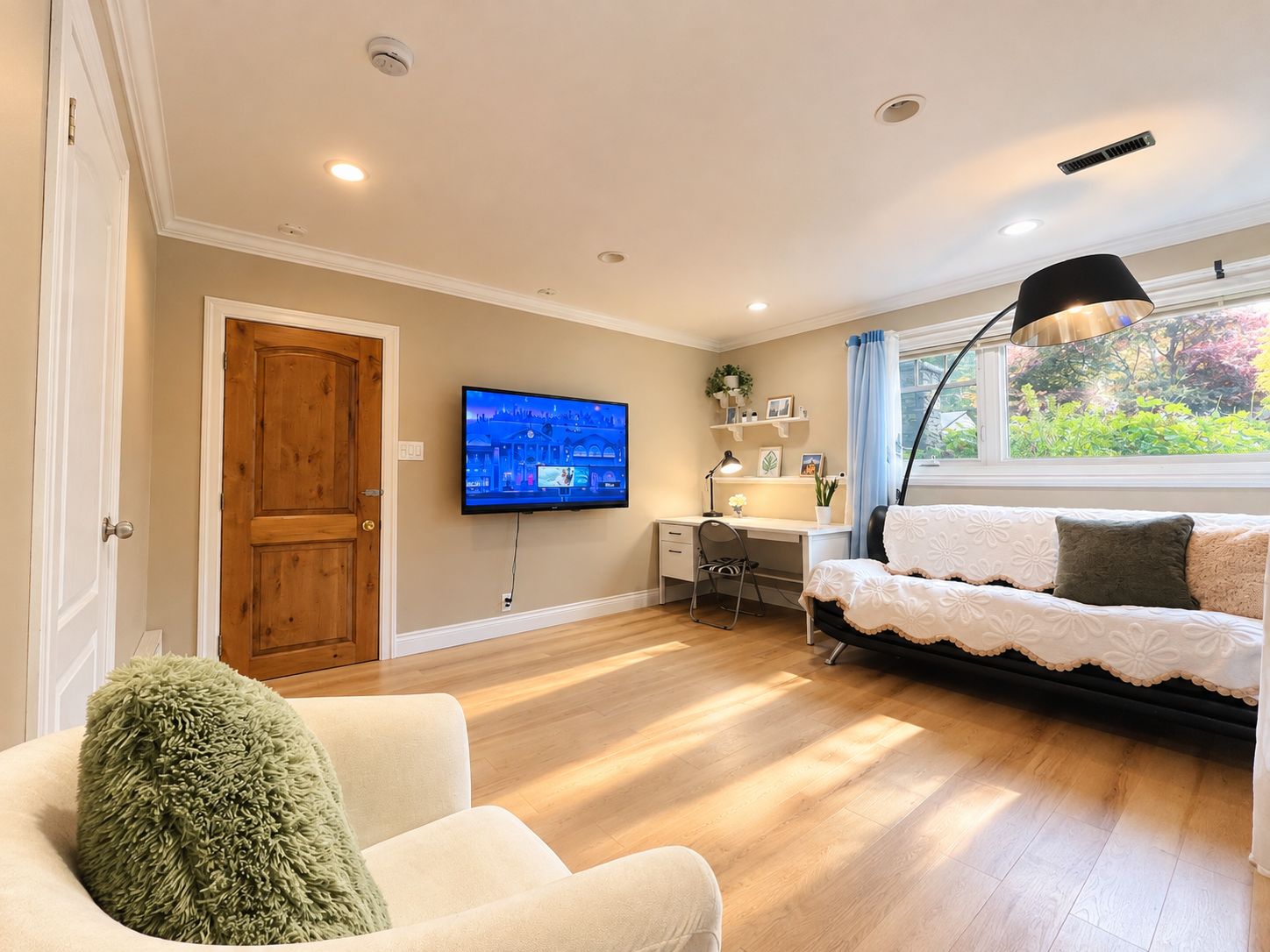 A living room with a television, a desk, a chair, a sofa, and a window with a view of greenery.
