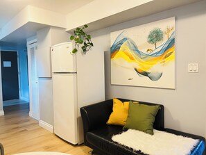 A living room with a white refrigerator, a black leather sofa with yellow and green cushions, a white furry throw, and a wall-mounted abstract painting.