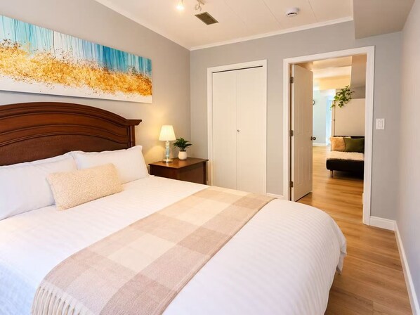 A hotel room with a large bed, a bedside table with a lamp, a painting on the wall, and a view into the living area.