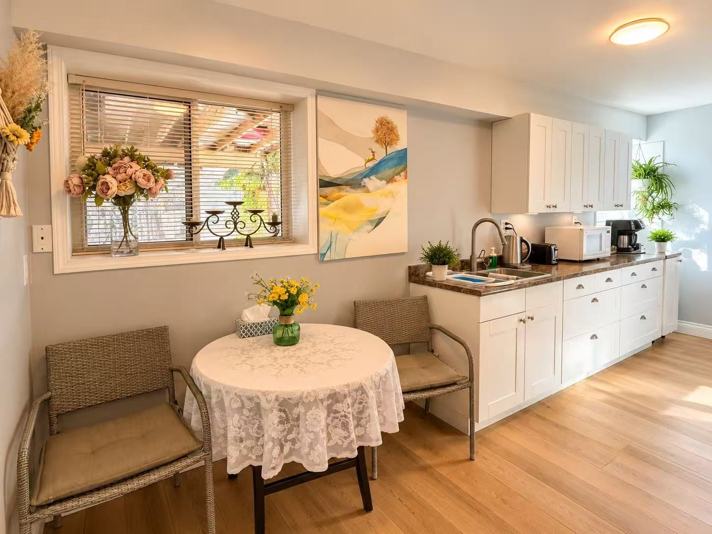 A modern kitchen with white cabinets, a dining table with a lace tablecloth, wicker chairs, and a window with blinds.