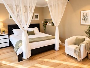 A bedroom with a four-poster bed, a bedside table with a lamp, a chair with a throw blanket, and a wall-mounted shelf with decorative items. - White Rock Suite + 3-min drive to the Beach (White Rock)