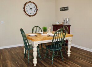 Un espace repas avec une table en bois, des chaises vertes, une horloge murale et un panneau indiquant l’emplacement du bar à café.