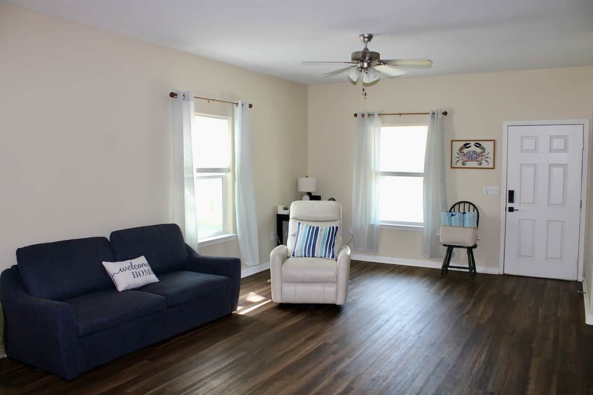 A living room with a dark blue sofa, a white armchair, and a small table with a lamp. There are two windows with white curtains, a white door, and a ceiling fan.