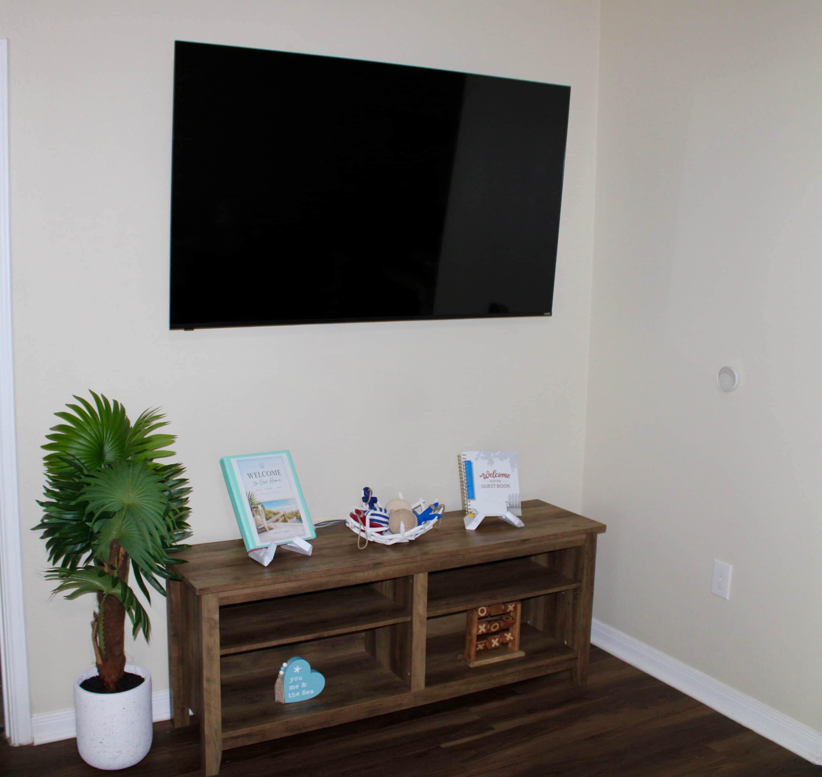 A room with a wooden console table, a potted plant, a welcome sign, a guest book, and a flat-screen TV mounted on the wall.