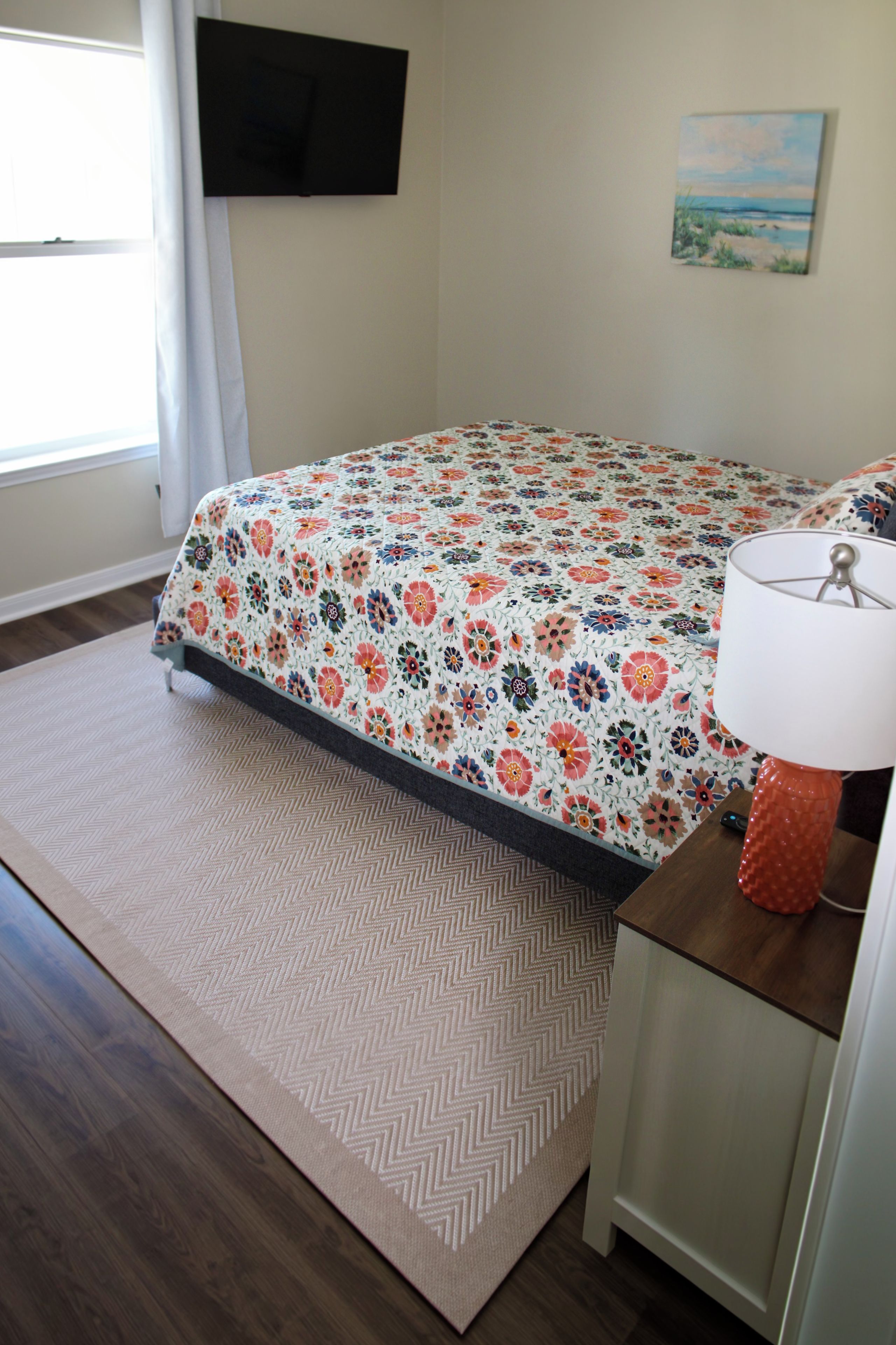 A bedroom with a bed, a nightstand, a lamp, a TV on the wall, and a window.