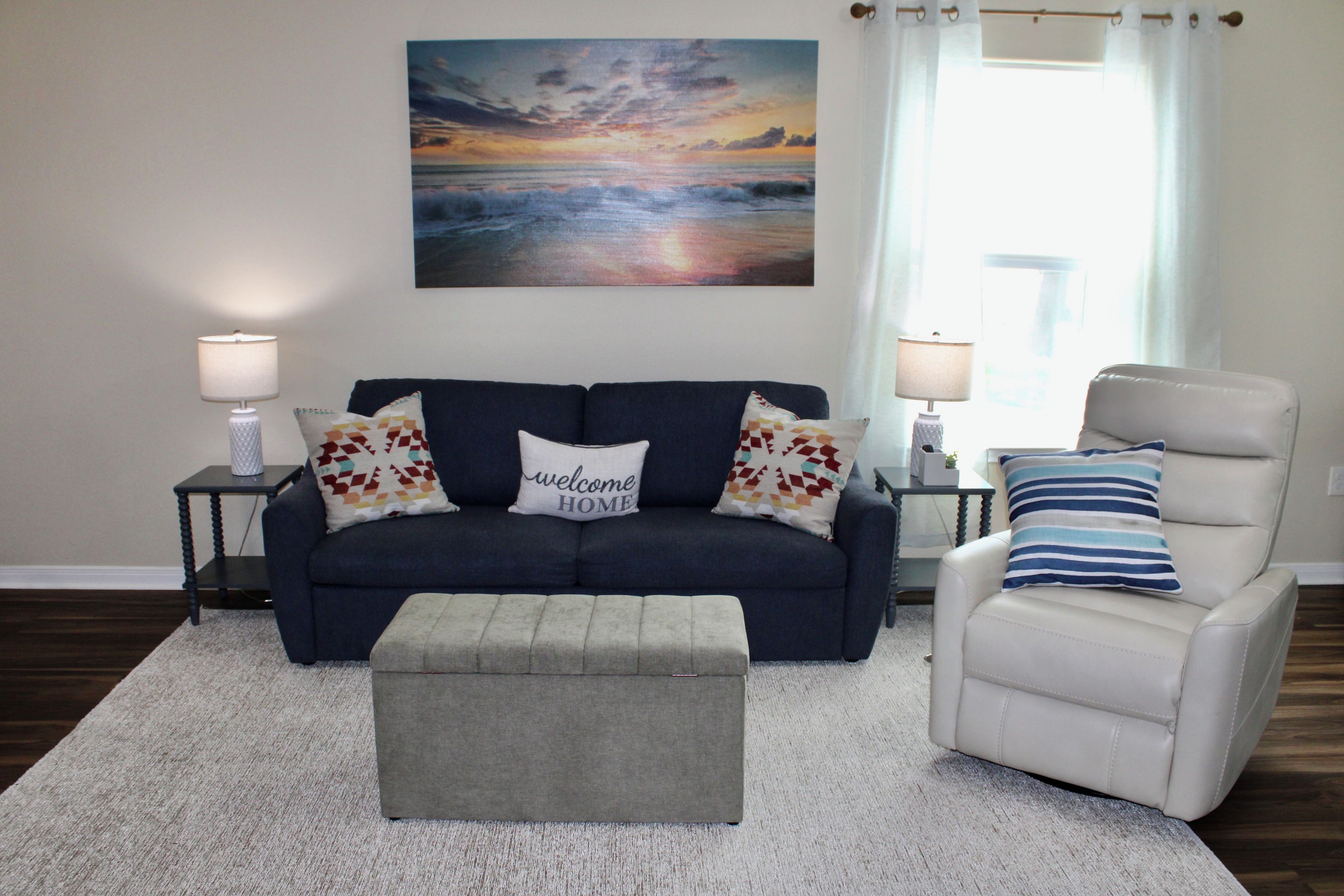 Uma sala de estar com um sofá azul escuro, uma poltrona branca, um banquinho cinza e um quadro de um pôr do sol sobre o oceano.
